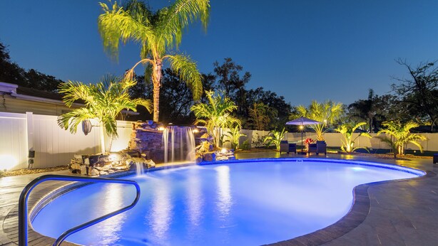 Una piscina con cascada, rodeada de palmeras y mobiliario exterior.
