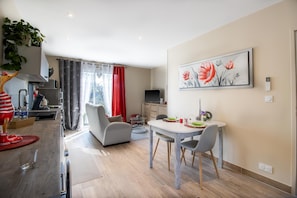 Een moderne woonkamer met een eethoek, een keuken met apparatuur, een groot raam met gordijnen, en een schilderij aan de muur.