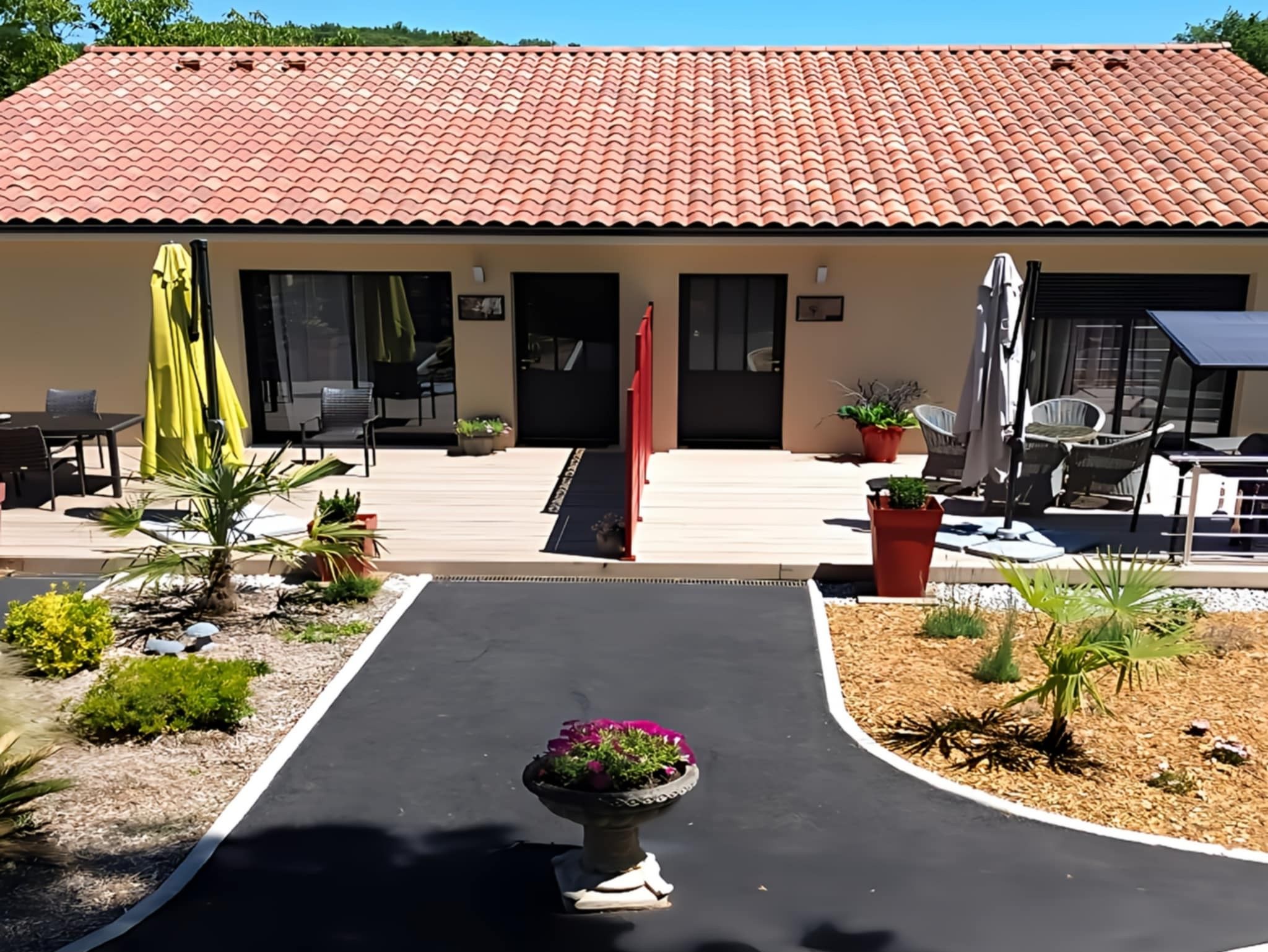 Un bâtiment avec un toit recouvert de tuiles rouges, un patio abrité avec des meubles d’extérieur et un jardin aménagé avec un parterre central.