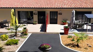 Un bâtiment avec un toit recouvert de tuiles rouges, un patio abrité avec des meubles d’extérieur et un jardin aménagé avec un parterre central.