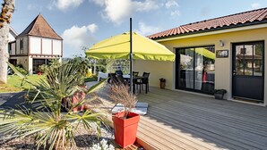 Eine Holzterrasse mit Gartenmöbeln, einem gelben Sonnenschirm und einem Haus mit Ziegeldach.