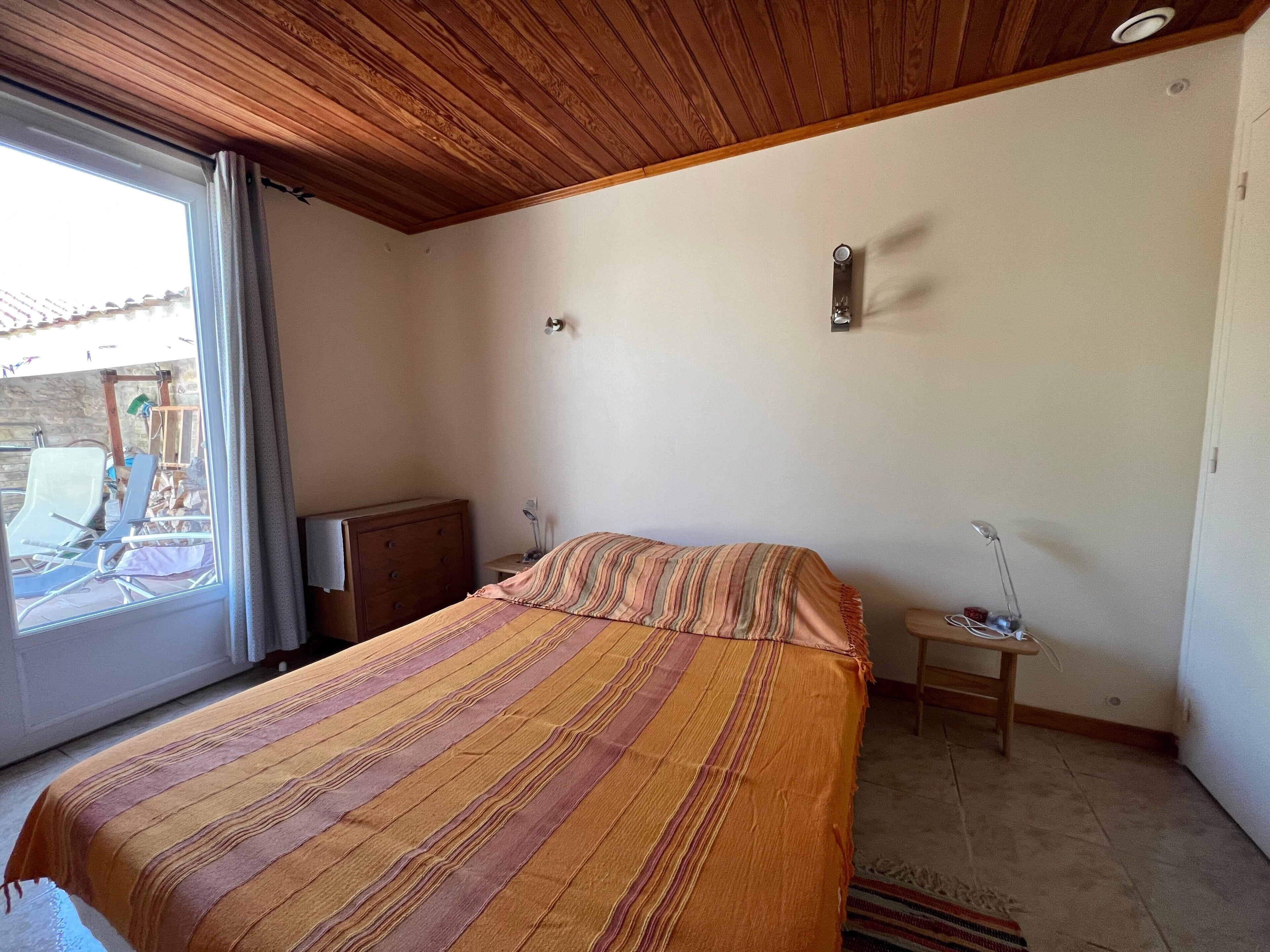 A bedroom with a bed, a nightstand, a lamp, a window, and a view of the outside.