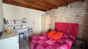 A bedroom with a stone wall, wooden ceiling beams, a bed with a pink and orange bedspread, a desk with books, and a small stool.