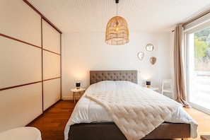 Une chambre à coucher avec un lit, un luminaire suspendu, une table de chevet et une vue sur l’extérieur.