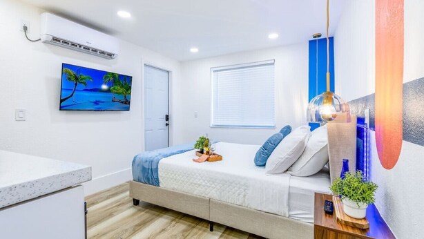 Un dormitorio moderno con cama, mesita de noche, aire acondicionado de pared, una ventana con persianas y un cuadro de una escena de playa.