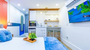 Una habitación de hotel moderna con una cama, una kitchenette pequeña, un televisor y un cuadro de una playa tropical.
