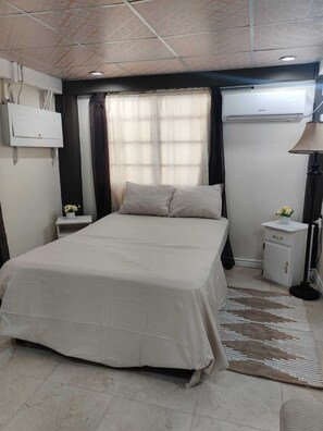 A bedroom with a large bed, a nightstand, a bedside table, a lamp, an air conditioner, and a window with curtains. - Mandarin Villa Vacation Package - Toco
(Inclusive of Both Villas) (Sangre Grande Regional Corporation)