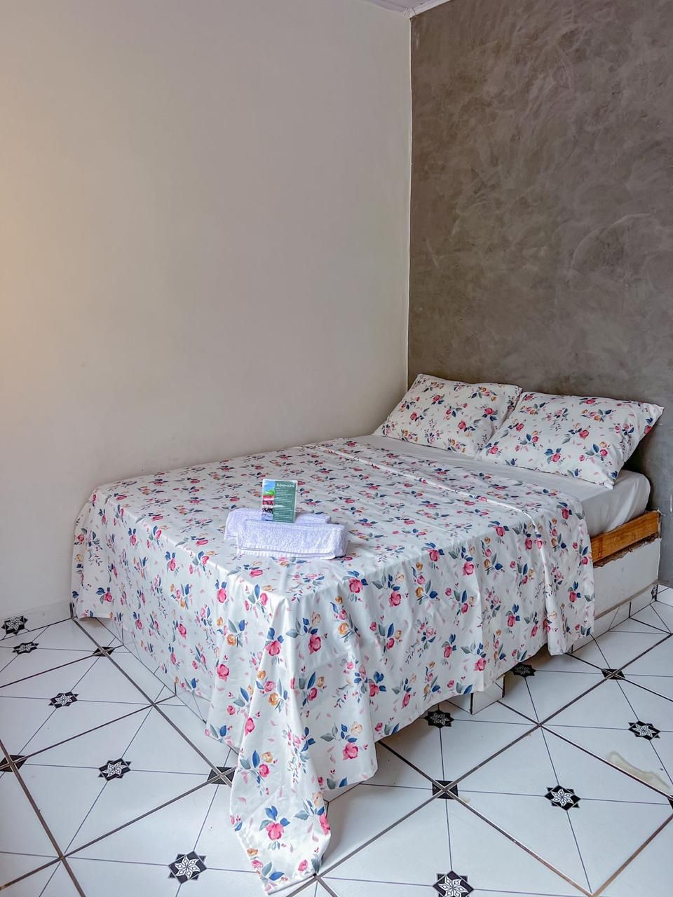A single bed with a floral bedspread, a small table with a folded cloth and a brochure, and a tiled floor with a geometric pattern.