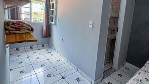 A room with a tiled floor, a bed with a floral bedspread, a wooden shelf with a blanket, a window with curtains, and a door leading to a bathroom. - Pousada Sempre Viva Lençóis (Lençóis)