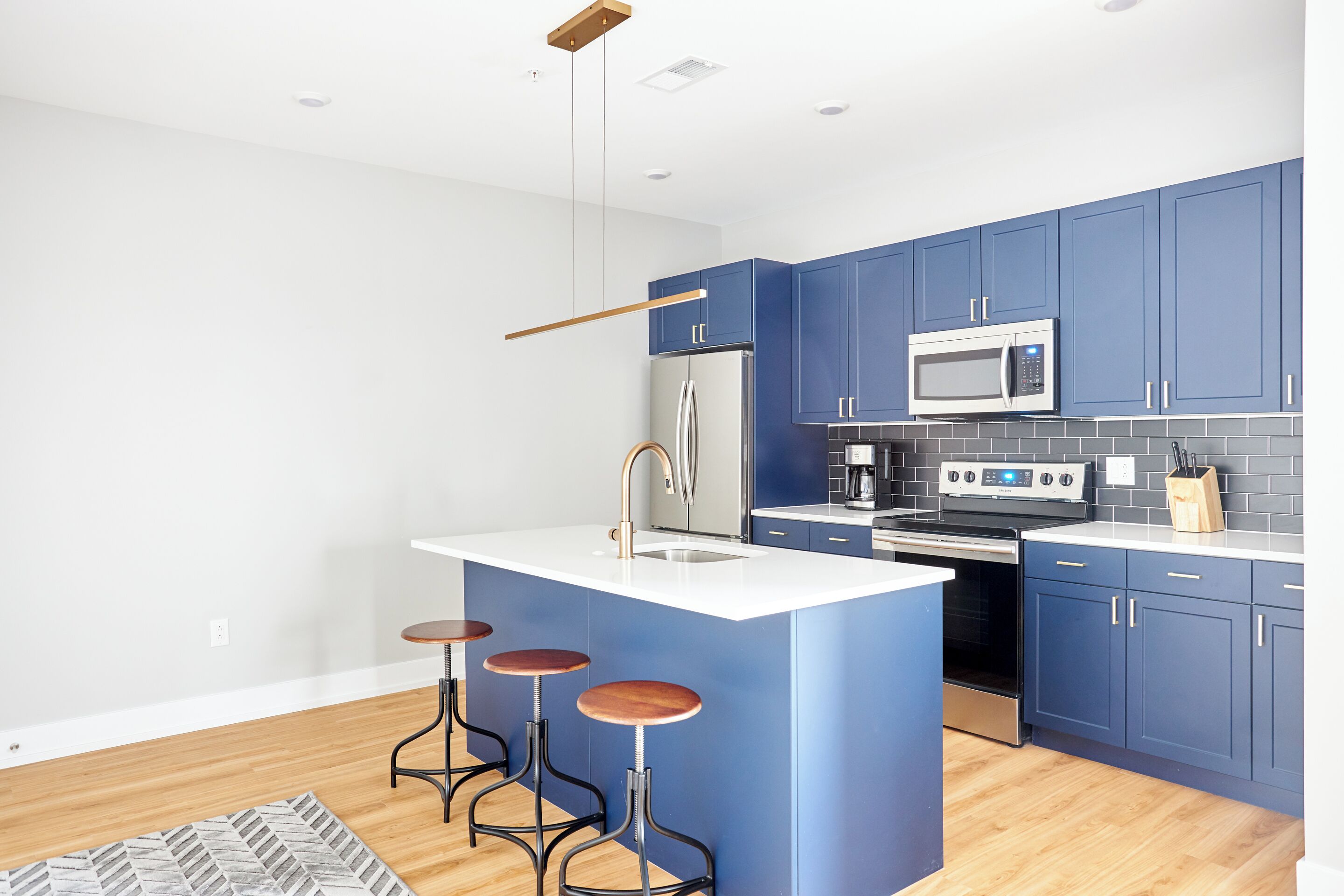 A modern kitchen with blue cabinets, a stainless steel refrigerator, a microwave, and a gas stove. There are three stools at the island.