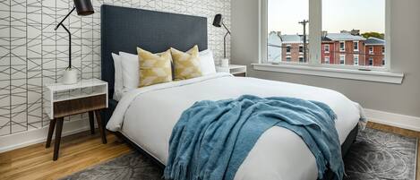 A bedroom with a bed, two bedside tables, a window with a view of buildings, and patterned wallpaper.