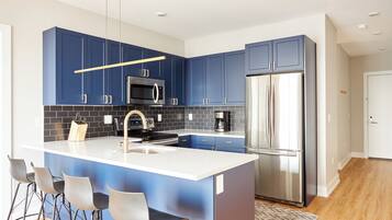 A modern kitchen with blue cabinets, a stainless steel refrigerator, a microwave, and a coffee maker. There are bar stools and a large island with a sink.