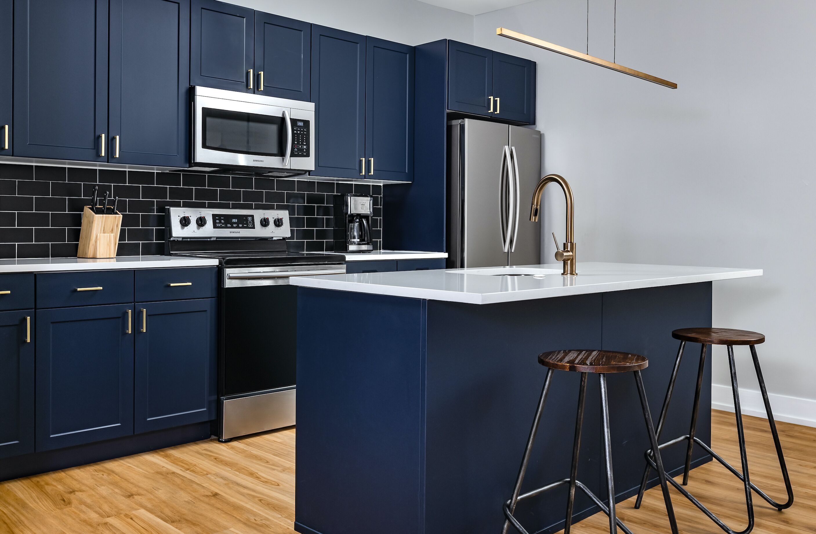 Una cucina moderna con mobili scuri blu, elettrodomestici in acciaio inossidabile, un piano di lavoro bianco e due sgabelli.