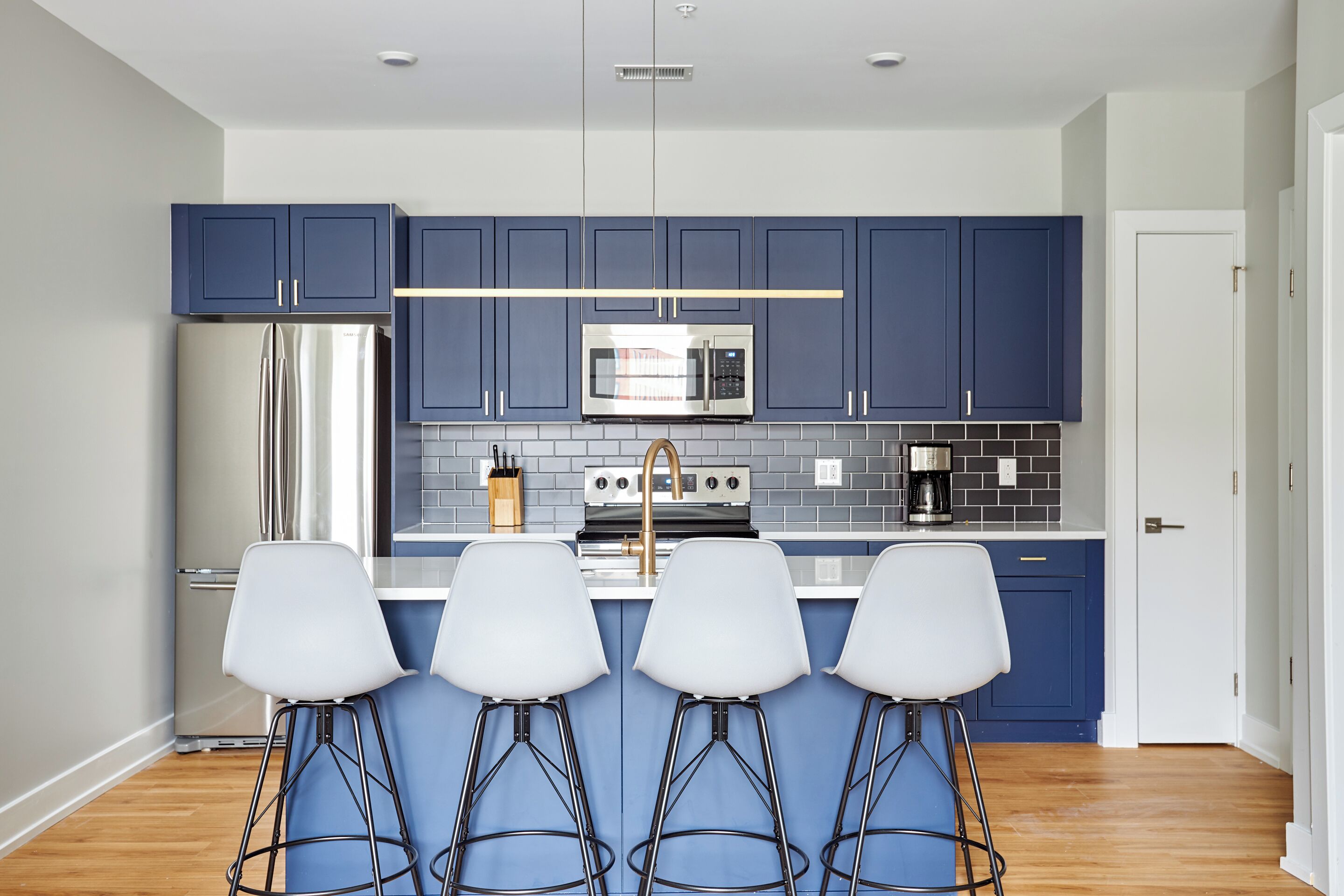 Eine moderne Küche mit blauen Schränken, einem Edelstahl-Kühlschrank, einer Mikrowelle und einer Kaffeemaschine. Drei weiße Barhocker sind vorhanden.