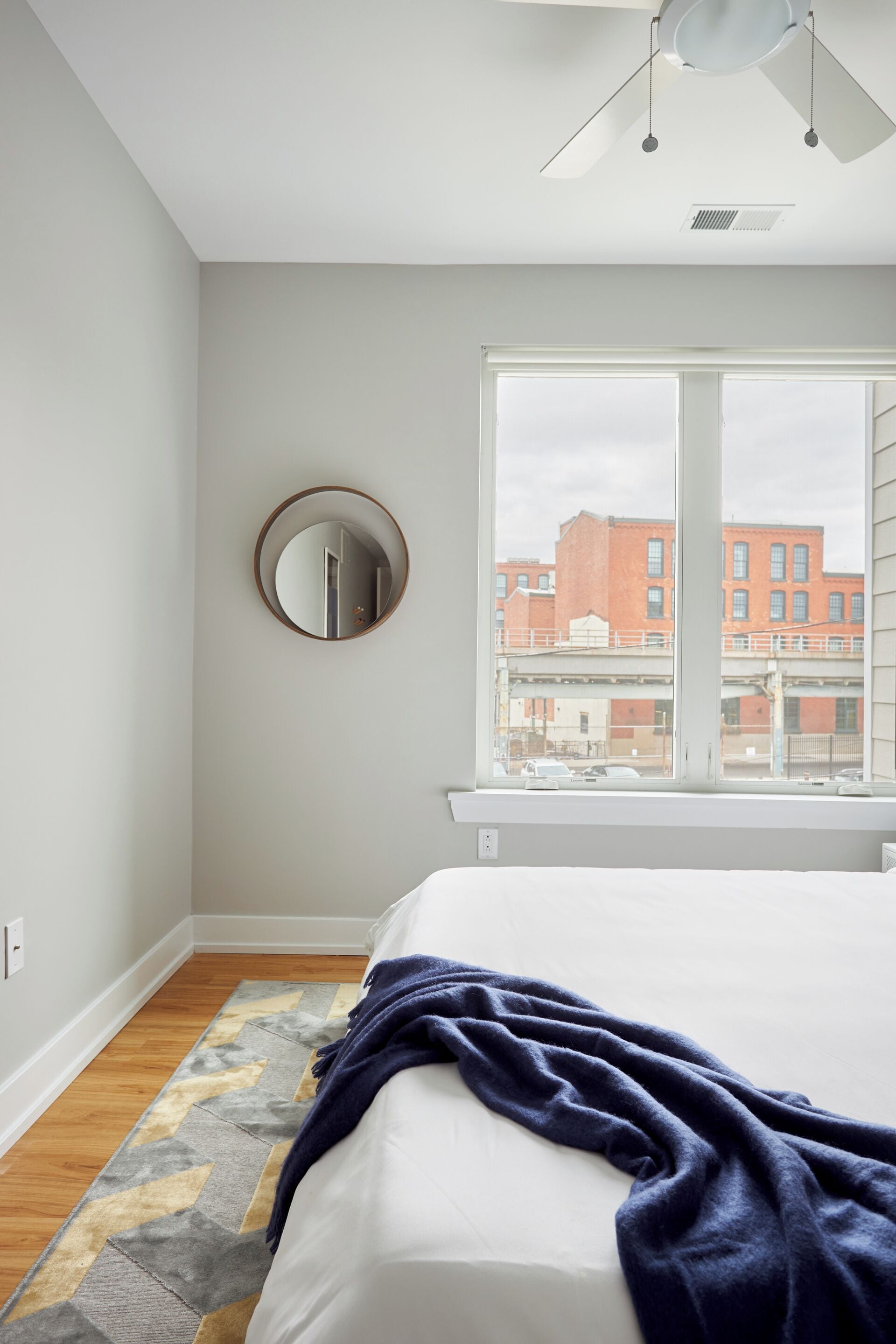 Une chambre à coucher avec un lit, une fenêtre donnant sur des immeubles, un miroir rond et un ventilateur de plafond.