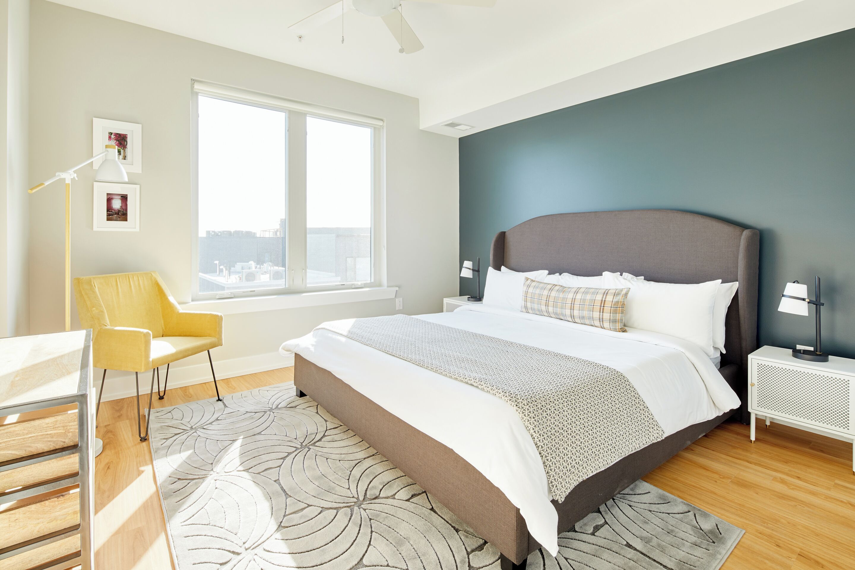 A modern bedroom with a large bed, a view of the city, a yellow chair, and a white desk.
