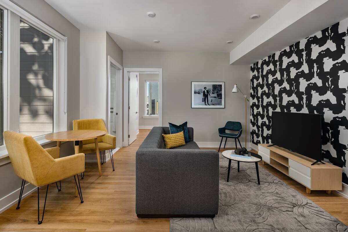 A modern living room with a grey sofa, yellow chairs, a small round table, and a TV on a wooden stand.