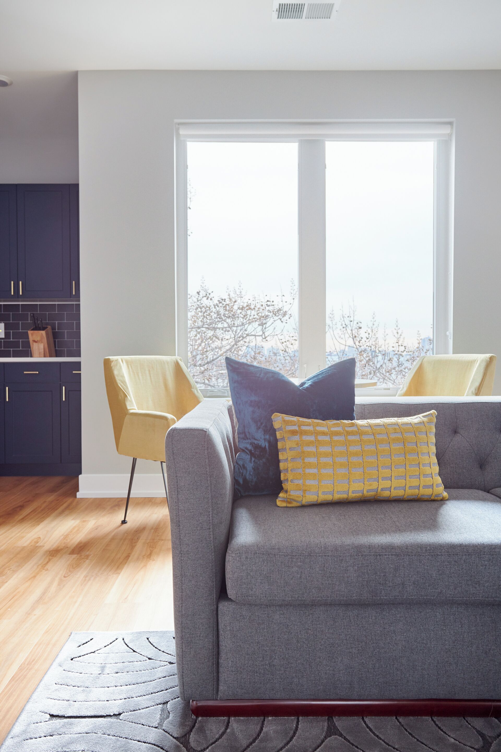 Ein modernes Wohnzimmer mit einem grauen Sofa, gelben und blauen Kissen und Blick ins Freie durch ein großes Fenster.