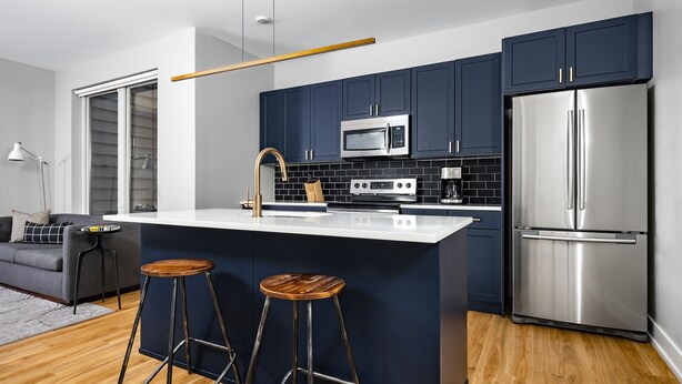 A modern kitchen with dark blue cabinets, stainless steel appliances, and a wooden bar stool. - StayCozy at The Frankford (Philadelphia)