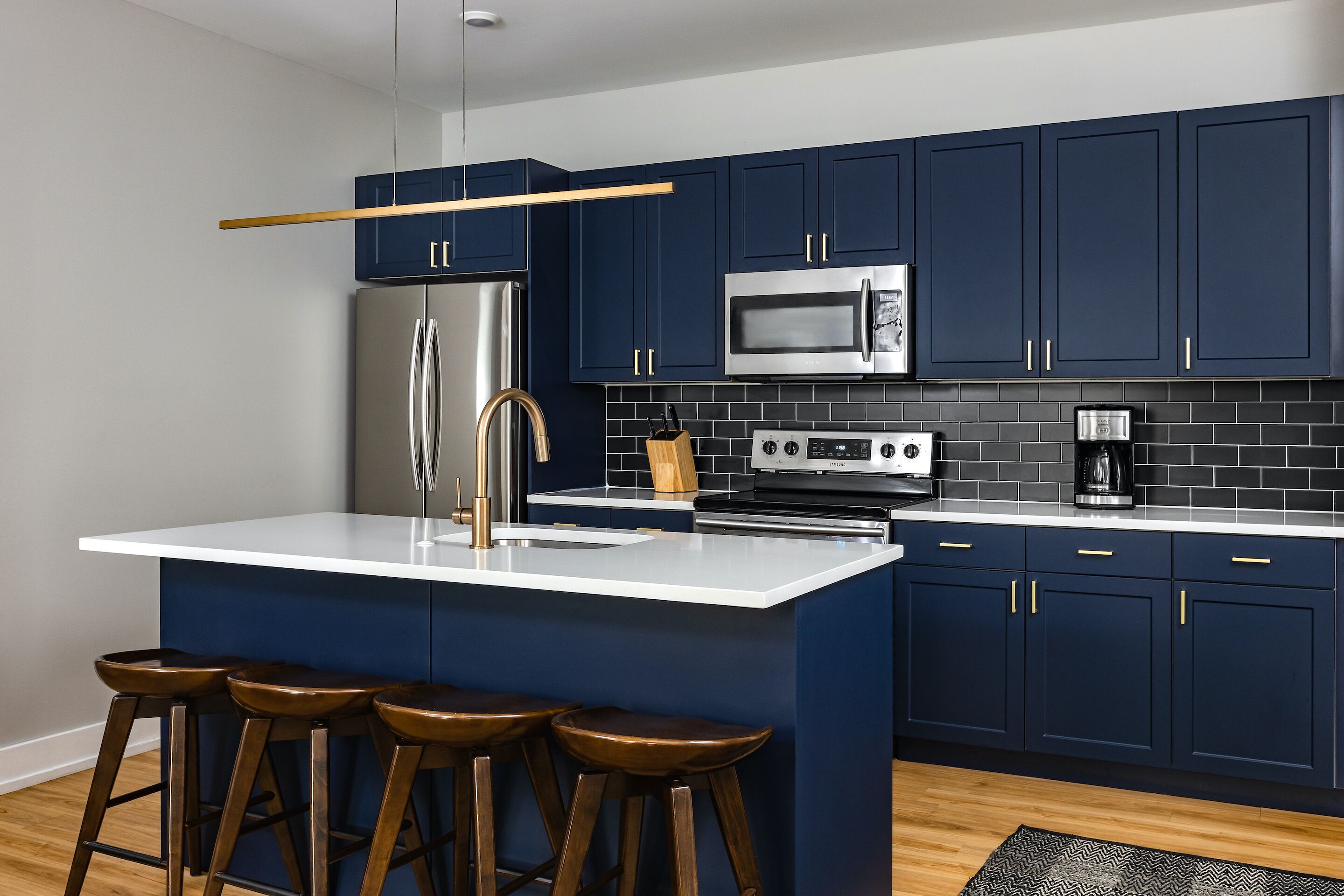 Een moderne keuken met donkerblauwe kastjes, roestvrijstalen apparatuur, een wit aanrecht en houten krukken.