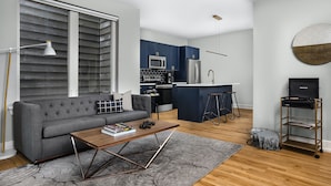 A modern living room with a grey sofa, a wooden coffee table, and a kitchen area with blue cabinets and stainless steel appliances. - StayCozy at The Frankford (Philadelphia)