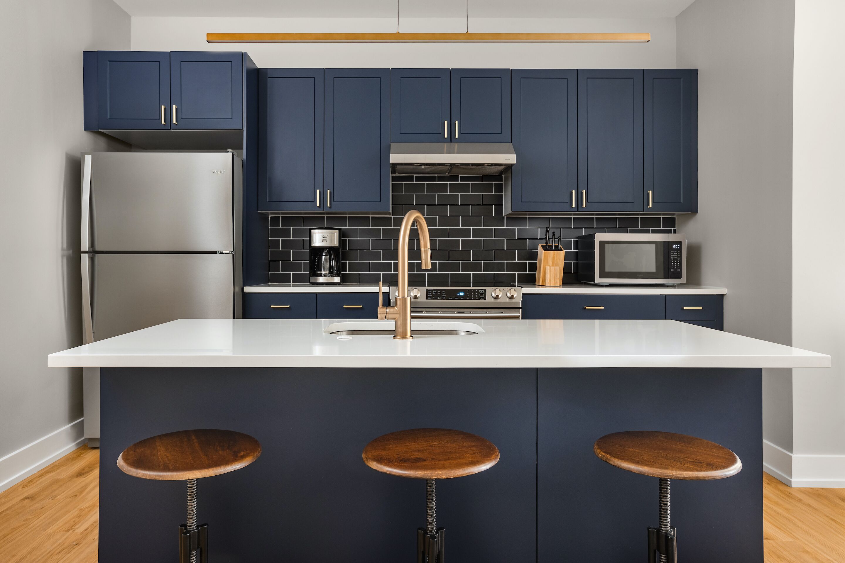 A modern kitchen with dark blue cabinets, a stainless steel refrigerator, a microwave, and a sink with a gold faucet. There are three wooden stools at the island.