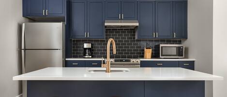 A modern kitchen with dark blue cabinets, a stainless steel refrigerator, a microwave, and a sink with a gold faucet. There are three wooden stools at the island.