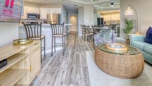 A modern kitchen with white cabinets, a microwave, and a refrigerator. There is a dining area with chairs and a table. The living room has a blue sofa and a round coffee table. - MyStay at 1102 Sea Crest - Sea Crest Resort Oceanfront Complex | Steps to Beach (Hilton Head Island)