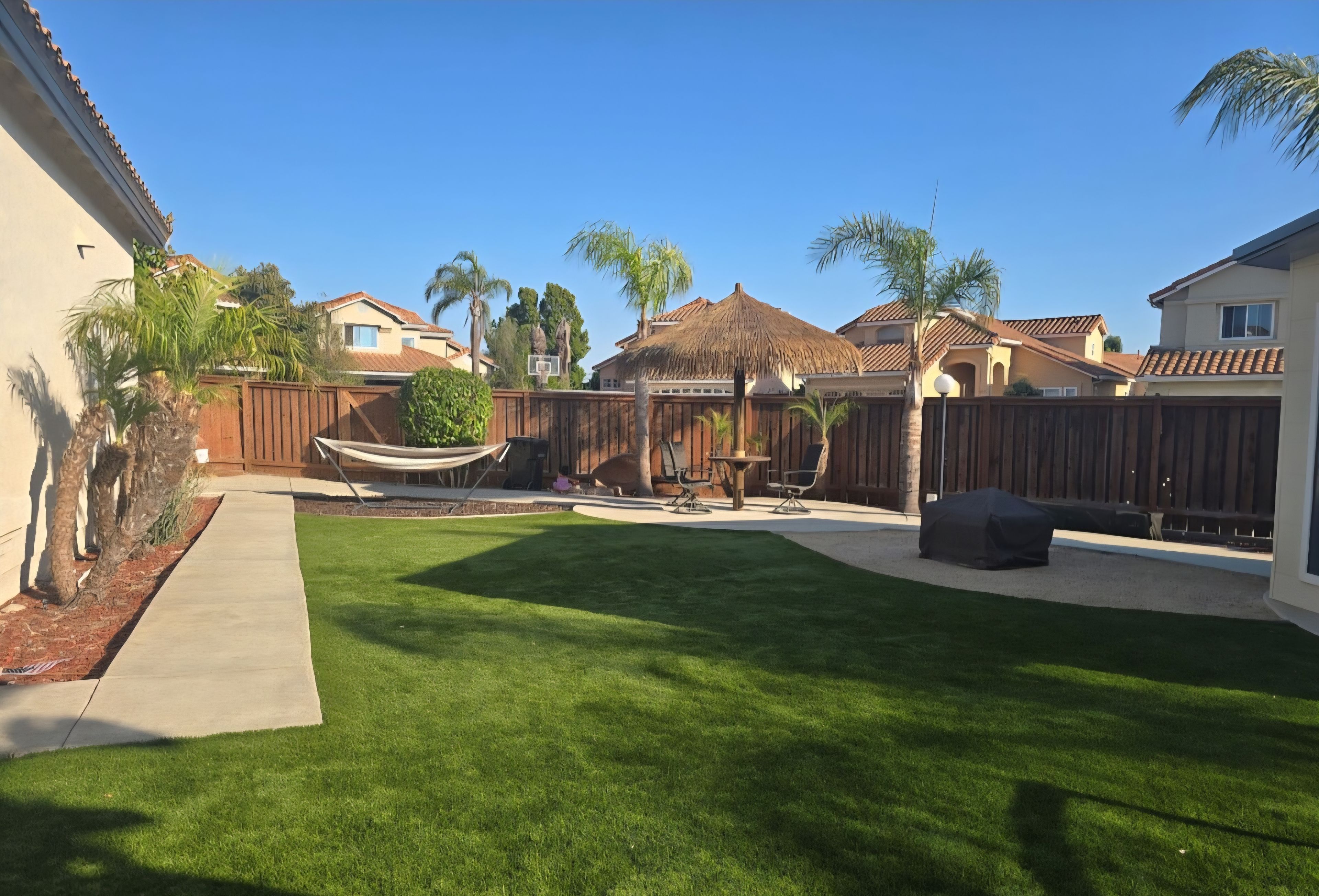 A backyard with a well-maintained lawn, a hammock, outdoor furniture, and a thatched-roof structure.