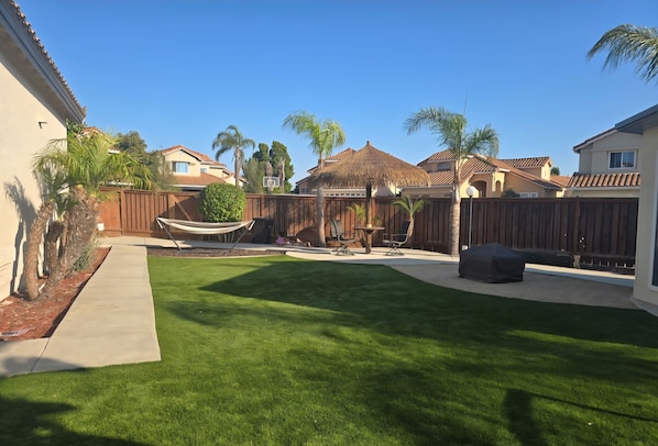 A backyard with a well-maintained lawn, a hammock, outdoor furniture, and a thatched-roof structure.