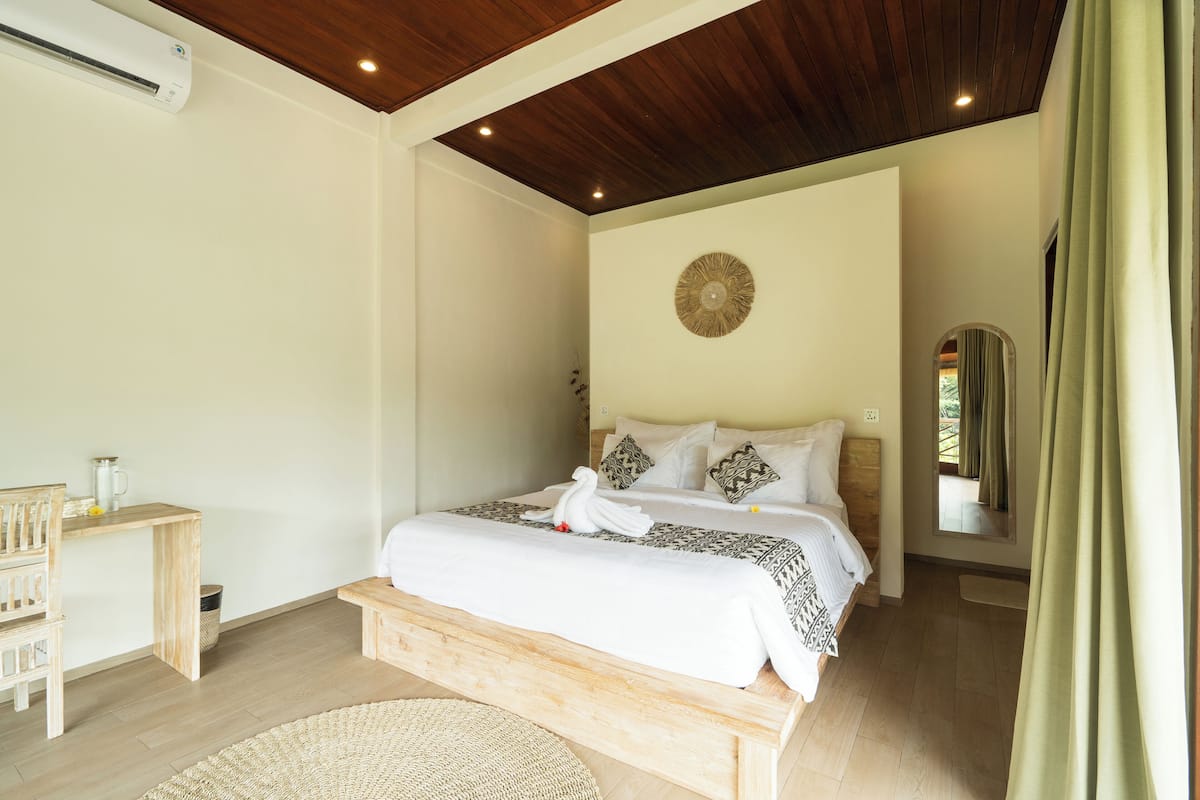 A bedroom with a wooden bed, a bedside table, a mirror, and a wall-mounted air conditioner.