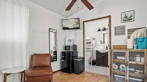 Una sala moderna con un sillón de cuero marrón, una mesa blanca, un televisor, un microondas, un refrigerador pequeño, un lavabo y un armario con utensilios de cocina.