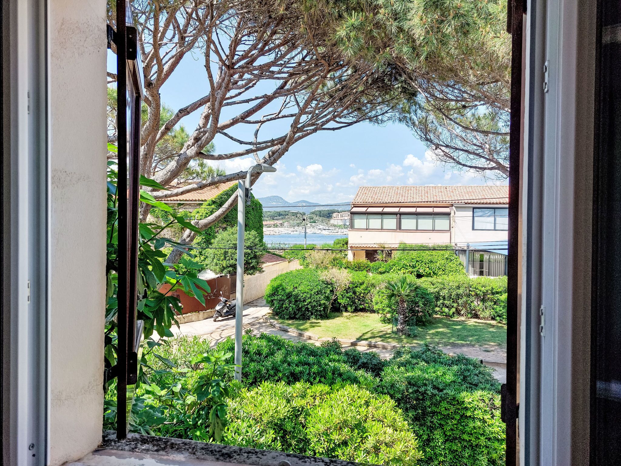 A view through a window of a garden with a paved area, a scooter, and a building with a tiled roof.