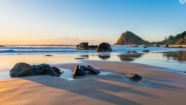 A beach with large rocks in the foreground and a hill in the background.