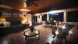 Una sala de estar acogedora con pisos de madera, ventilador de techo, sofá, mesa de centro con flores y un comedor con sillas y mesa.