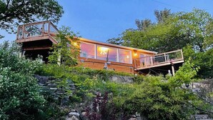 Una cabaña de madera moderna con ventanas amplias, un balcón y un cimiento de piedra rodeada de abundante vegetación.
