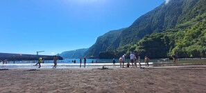 Uma praia com pessoas caminhando e nadando, cercada por colinas verdes exuberantes.