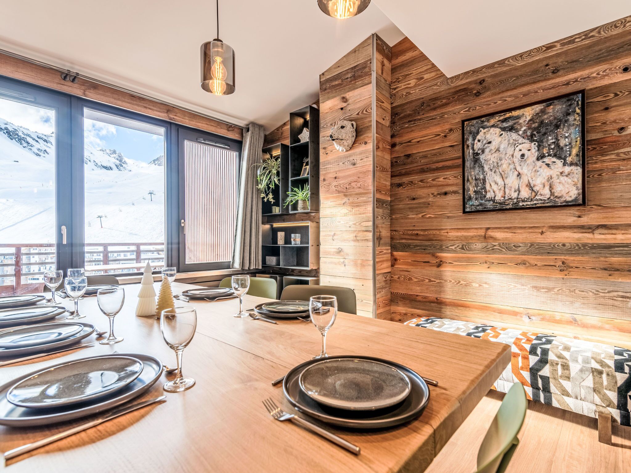 Un coin repas avec une table en bois dressée pour quatre personnes, de grandes fenêtres offrant une vue sur les montagnes et un mur rustique orné d’un tableau représentant un ours polaire.