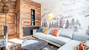 A modern living room with a wooden accent wall, a sofa with patterned cushions, a round coffee table, and a mountain mural.