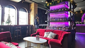 Eine Bar mit einer roten Couch, einem weißen Tisch und einer Theke voller Flaschen. Große Fenster und ein Weihnachtsbaum sind zu sehen.