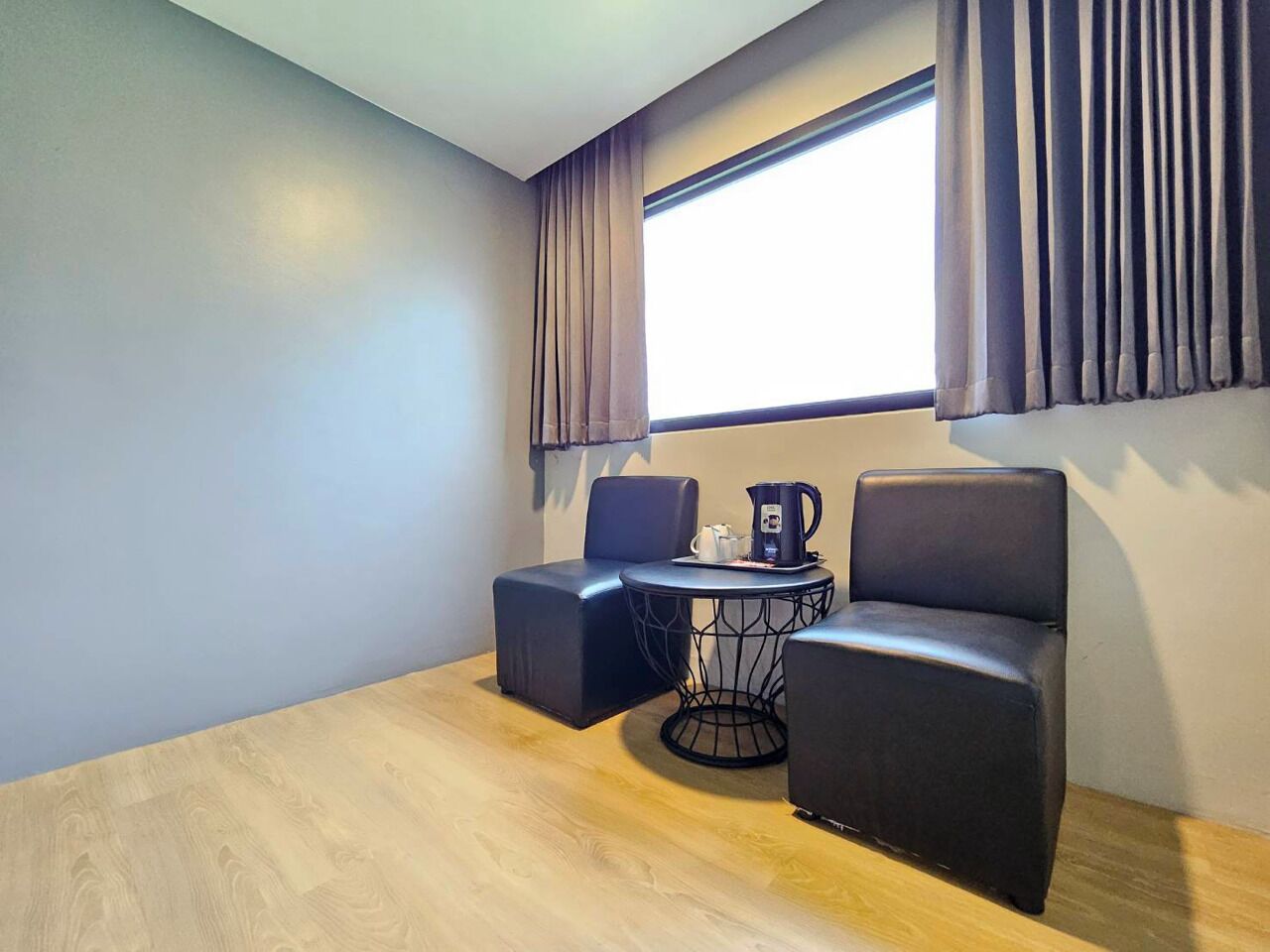 A modern room with a wooden floor, two black sofas, a small round table with a coffee pot, and a large window with curtains.