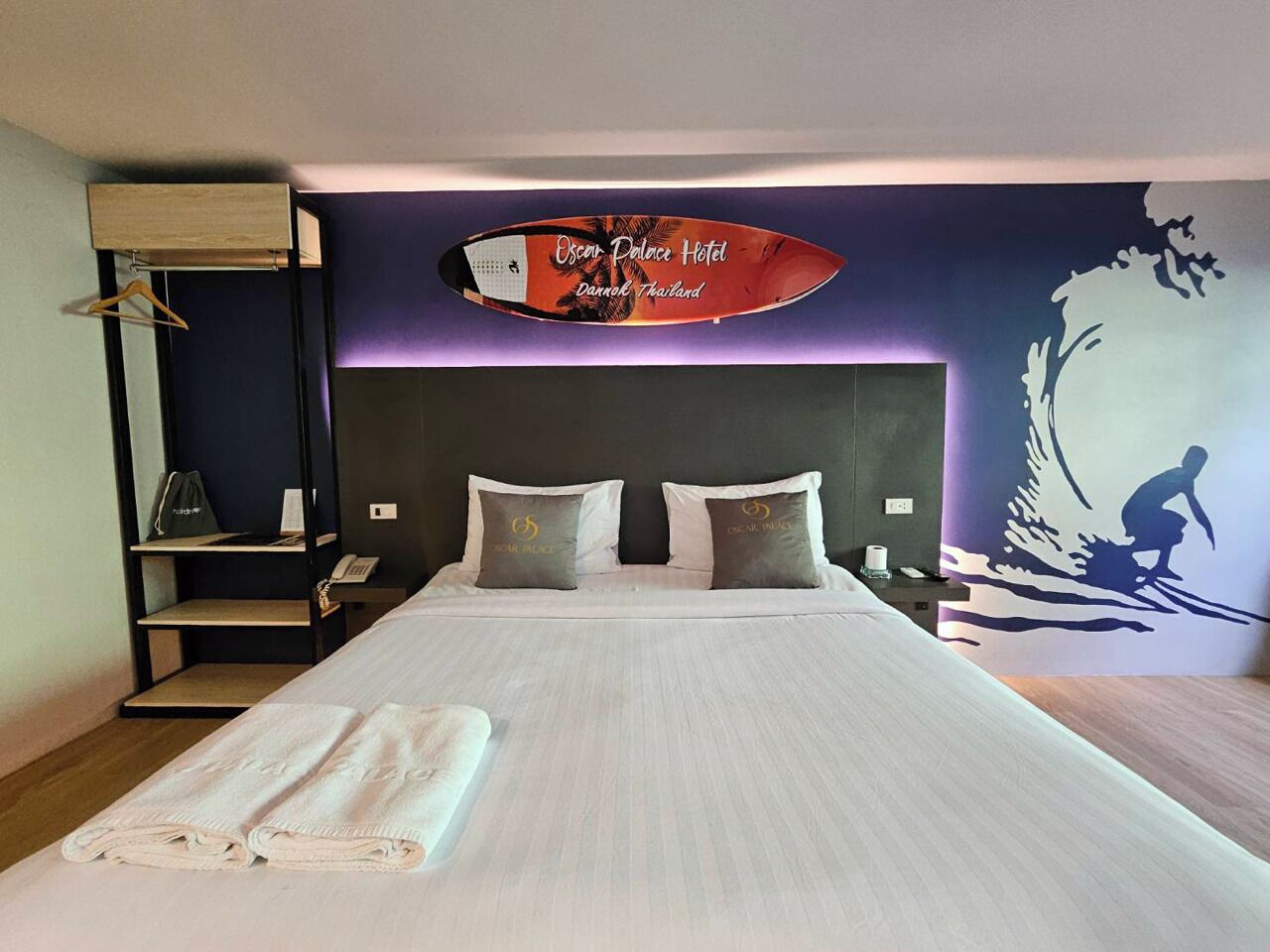 Habitación de hotel con cama, almohadas, una toalla, un estante y una pared decorada con una tabla de surf y texto.