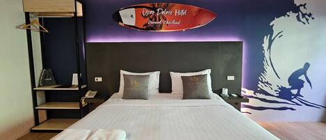 Habitación de hotel con cama, almohadas, una toalla, un estante y una pared decorada con una tabla de surf y texto.