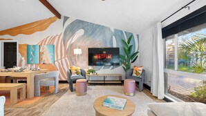 A modern living room with a large flat-screen TV, a dining area with a wooden table and chairs, and a large window with a view of greenery. - Beach Oasis: Hot tub, Pool, Mini-golf, Game Room (Seminole)