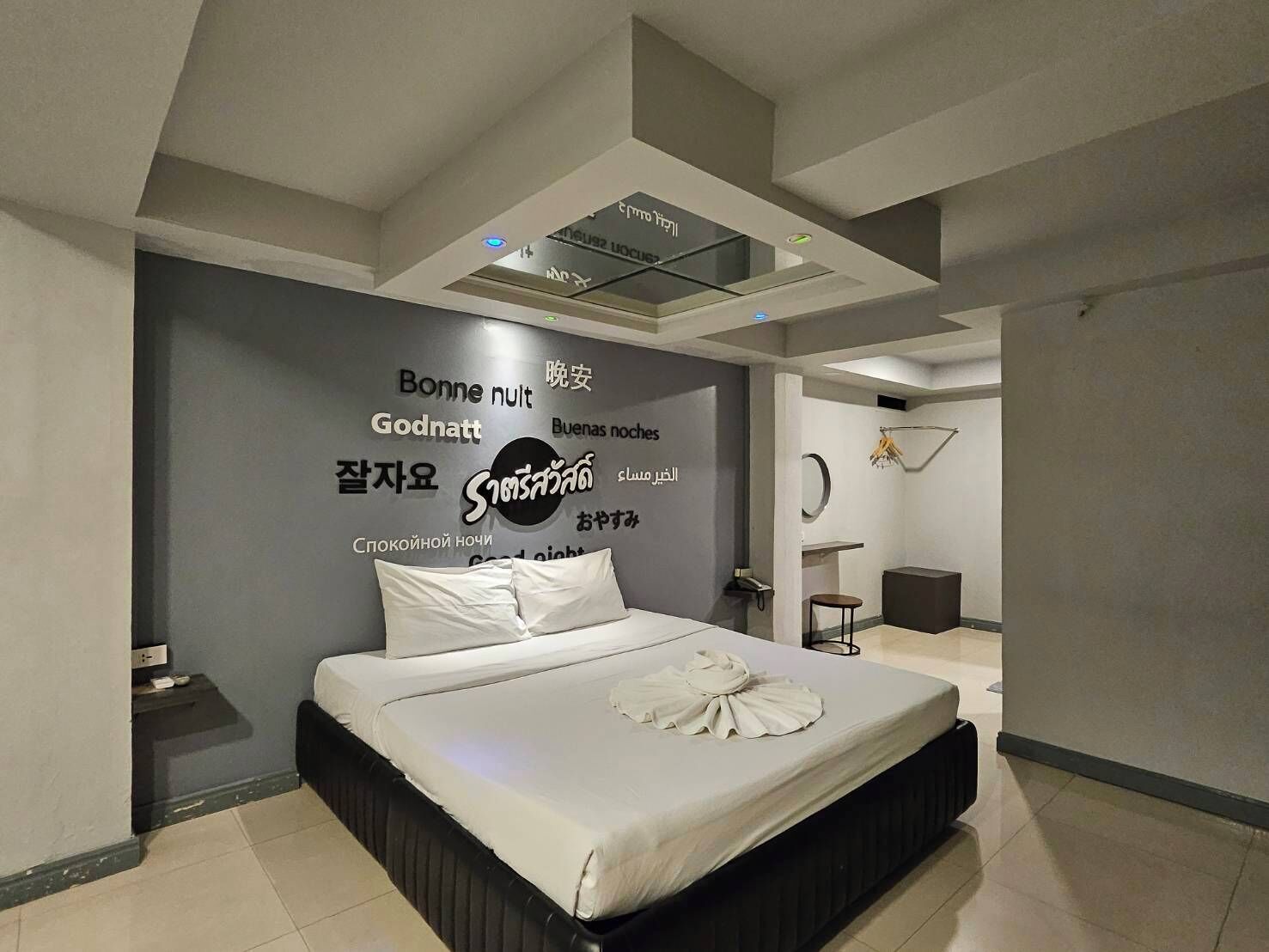 A modern bedroom with a bed, a wall with multilingual text, a ceiling light, and a small table with a chair.