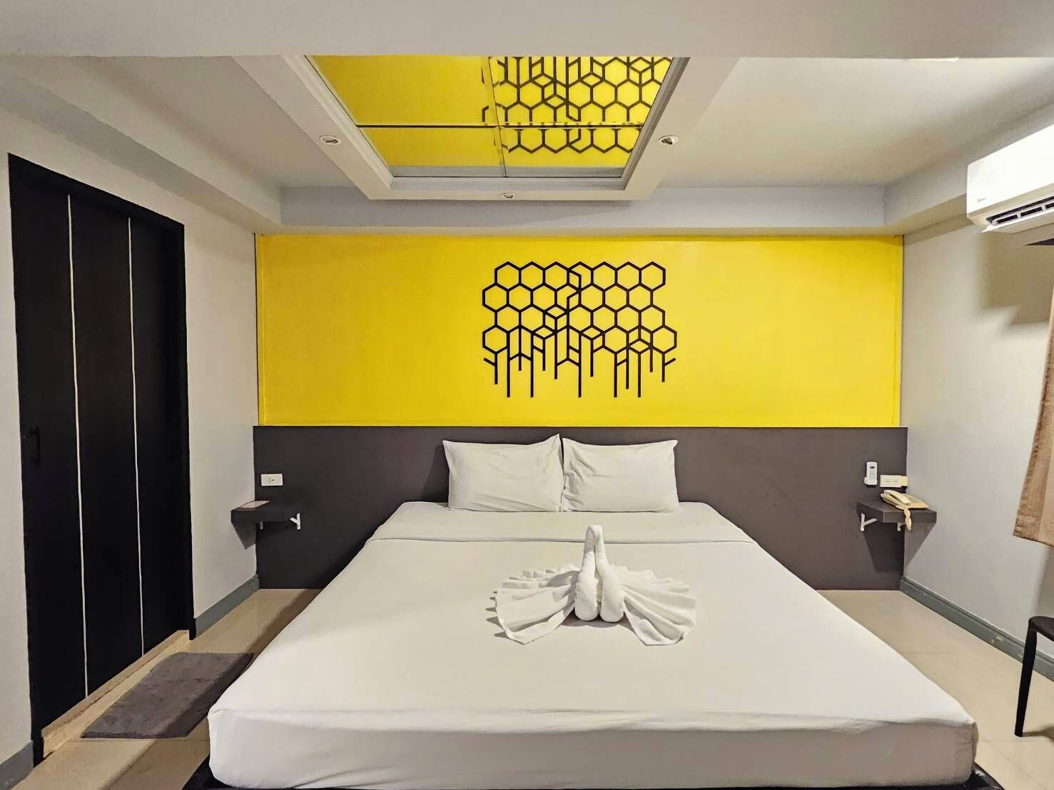 Ein Hotelzimmer mit einem großen Bett, einer gelben Wand mit Bienenwabenmuster, einem Oberlicht und einem schwarzen Kleiderschrank.