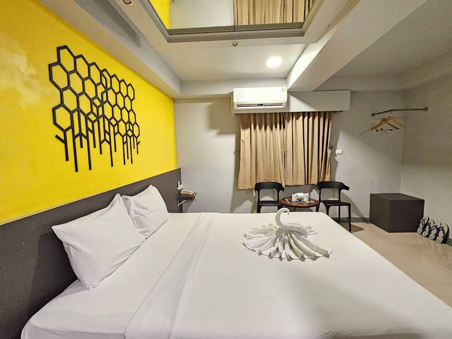 Habitación de hotel con cama, pared amarilla con diseño geométrico, zona de comedor con sillas y un equipo de aire acondicionado.