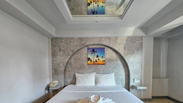 A hotel room with a large bed, a curved headboard, a painting of palm trees, and a skylight. - Hollywood Dannok Hotel (Sadao)