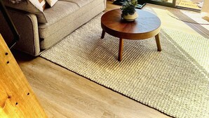 Una sala de estar acogedora con techo de madera, un sofá con cojines, una mesita de centro y vistas a una terraza y árboles a través de amplios ventanales.