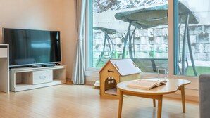 Een moderne woonkamer met een flatscreen televisie op een houten standaard, een klein houten tafeltje met een boek, en een huisje als speelhuisje.